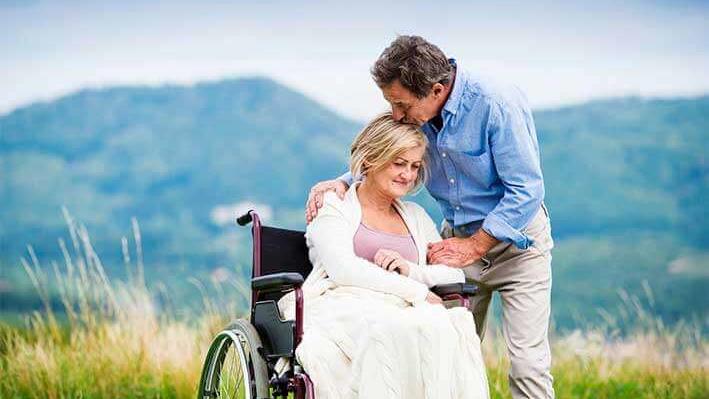 Man lightly hugs woman seated in wheelchair outdoors and kisses her head.