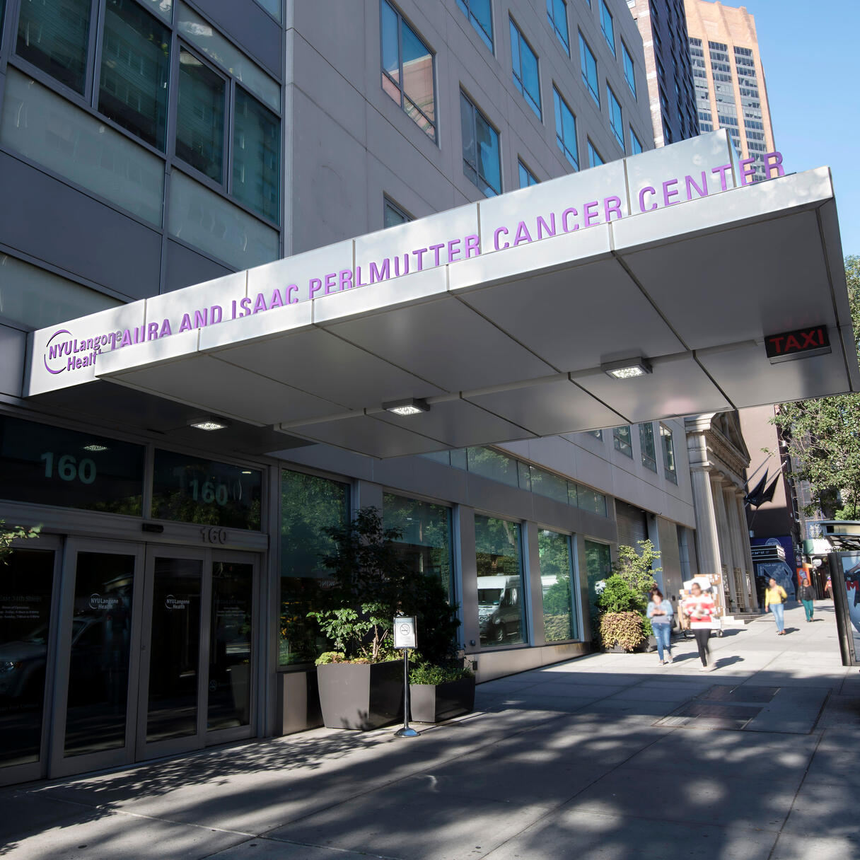 An exterior photo of the Laura and Isaac Perlmutter Cancer Center at NYU Langone Health. Only the lower floors of the building are visible along with the awning of the building that contains a sign with the name of the center. In the background, other tall buildings and trees can be seen along with cars on a street and people walking on a sidewalk. The sky is blue and cloudless.