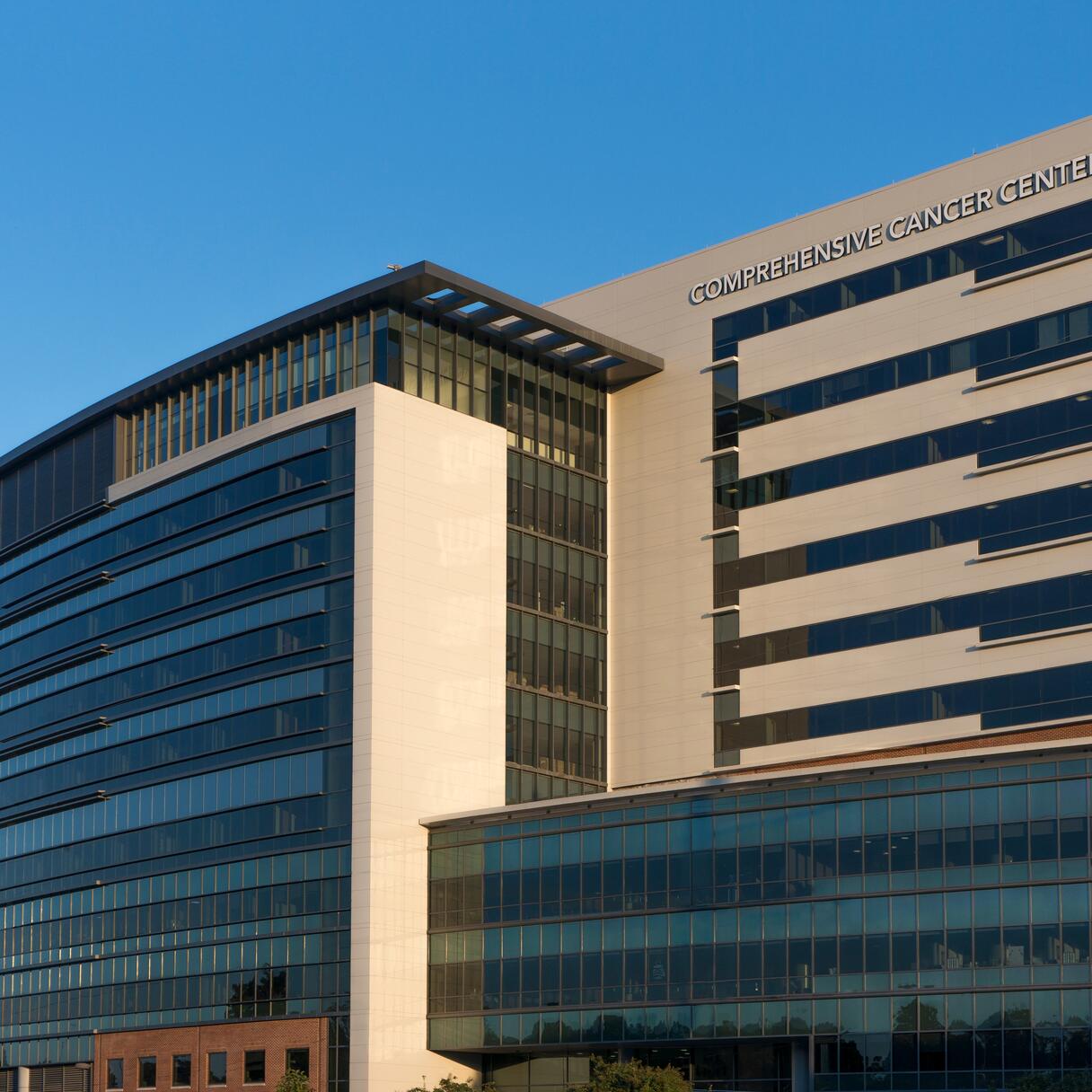 Exterior view of Wake Forest Baptist Comprehensive Cancer Center (WFBCCC). The building is a modern, multi-story building labeled "Comprehensive Cancer Center" and features large glass windows and a sleek, curved design, reflecting the sunlight.