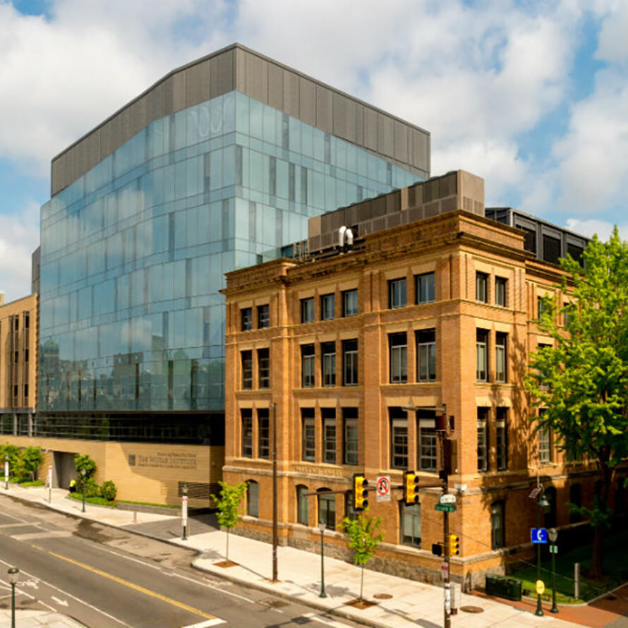 Modern architecture of The Wistar Institute featuring a striking glass cube addition connected to a historic brick building. The contemporary blue-glass structure rises several stories above the original brownstone research facility.