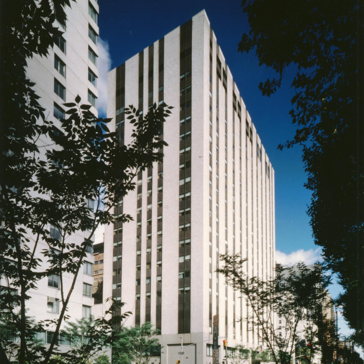 External view of Memorial Sloan Kettering Cancer Center. The building has a modern design, featuring vertical white and dark panels. The structure is set against a clear blue sky, framed by trees and a street lined with parked cars.