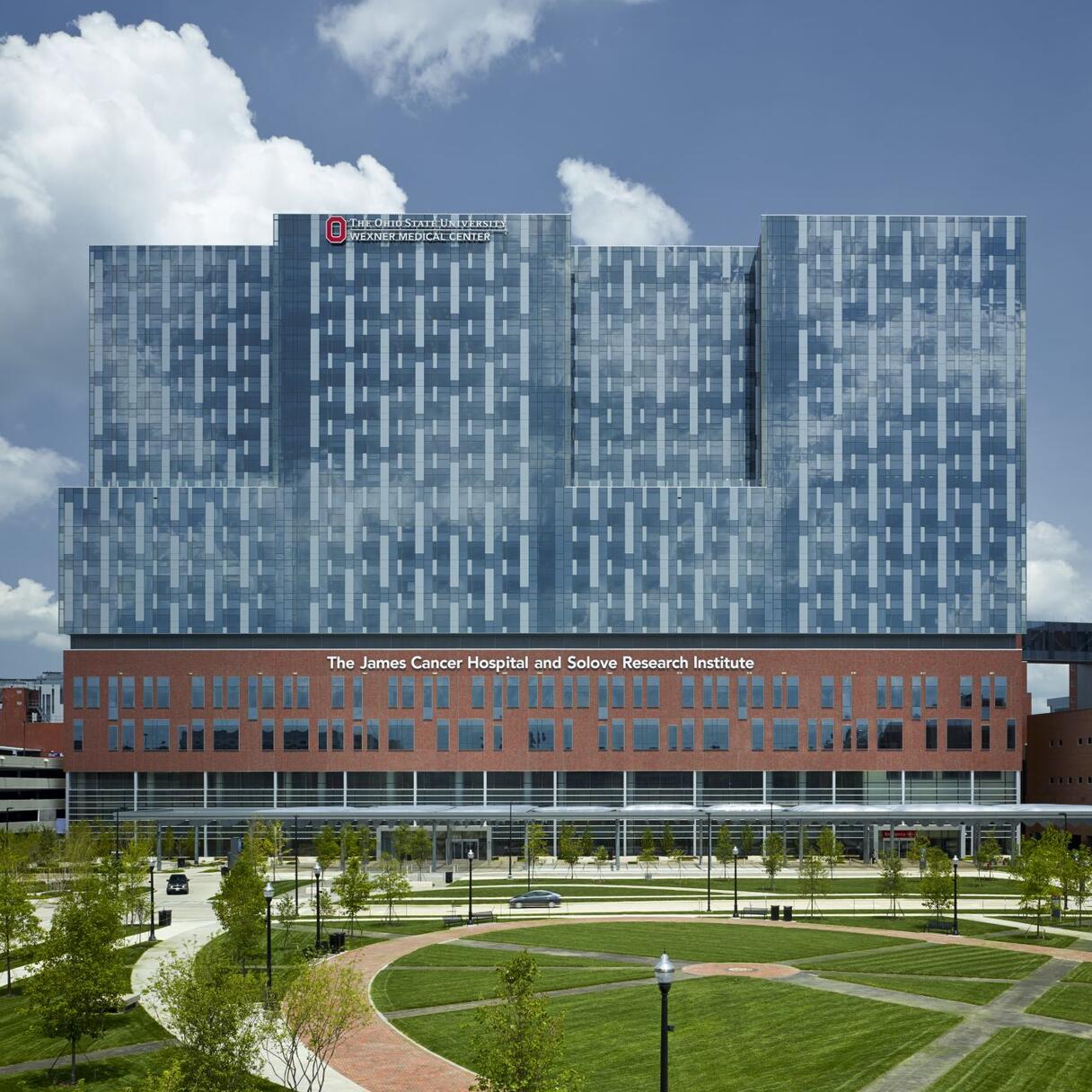 An exterior photo of the Ohio State University Comprehensive Cancer Center. The building is modern and mostly glass for the top 2/3s; the bottom is brown with windows. Smaller buildings can be seen on the left and the right; in front of the cancer center, a large green park with trees can be seen. The sky is blue with large fluffy clouds.