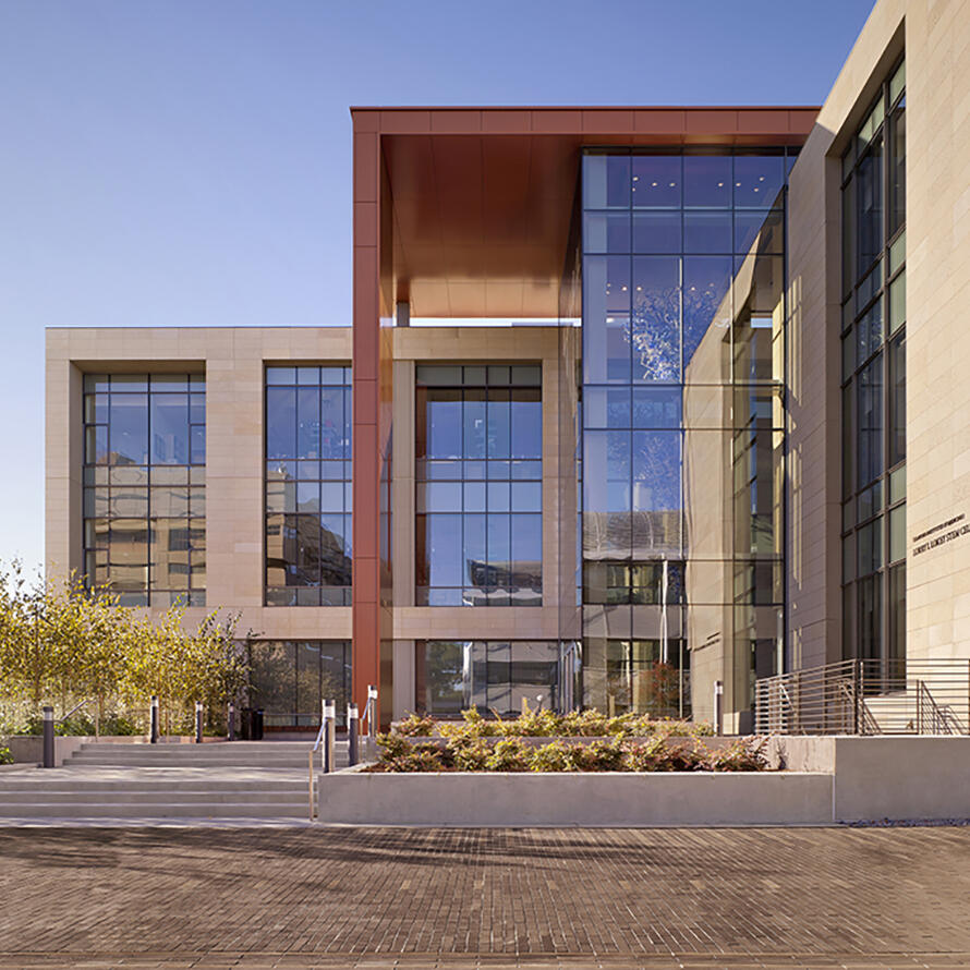 An exterior photo of the Stanford Cancer Institute. The building has hard edges and large glass panel windows that take up a majority of the exterior. The parts of the building that aren’t glass are greys and warm oranges. There are steps seen on both the left and right of the building, along with trees and bushes throughout. The sky is blue and cloudless. 
