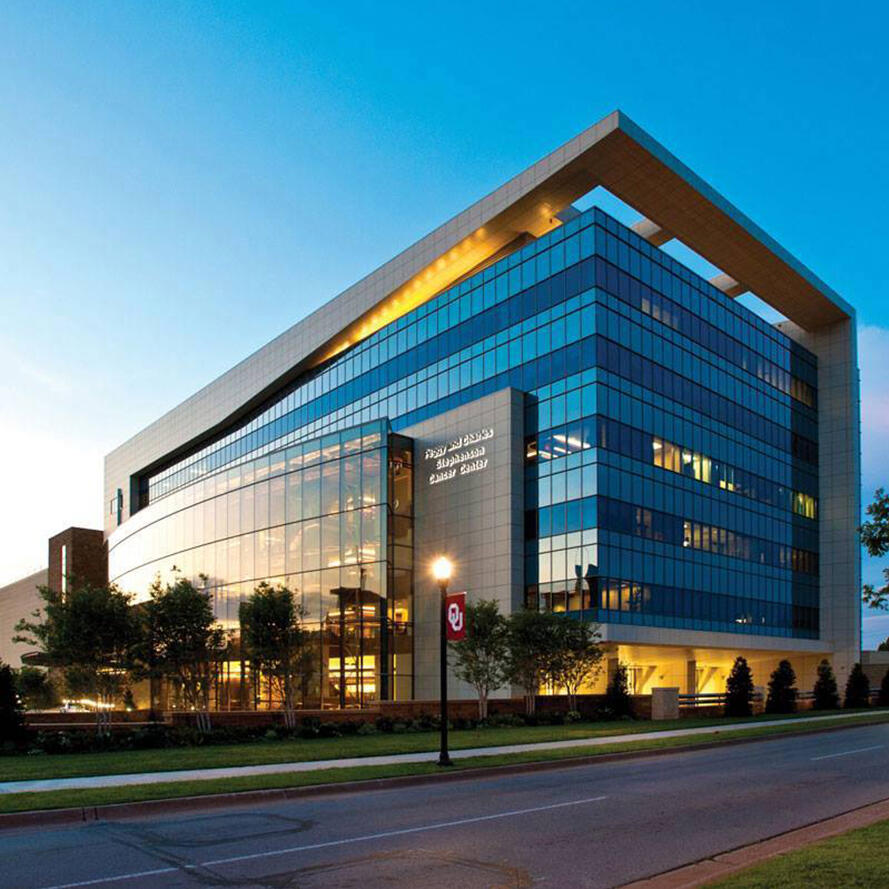 An exterior photo of the Stephenson Cancer Center. It is a modern building that is mostly made of glass windows. A large angular piece of the building at the top frames the lower floors. A row of trees runs along the length of the building. The sky is blue and the light hits the building’s windows in a way to suggest sunrise or sunset.