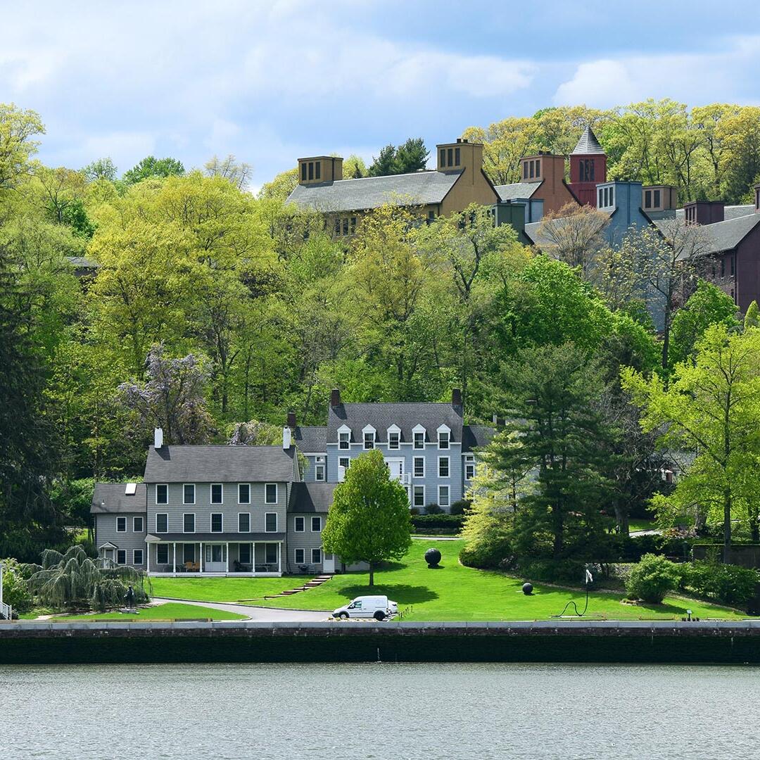 Cold Spring Harbor Laboratory Cancer Center buildings on a green hillside overlooking a waterfront, surrounded by dense trees under a partly cloudy sky.