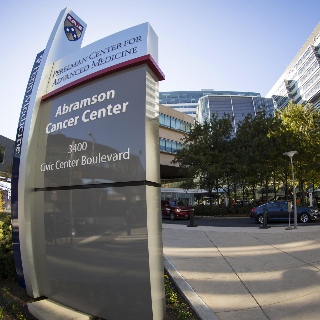 Abramson Cancer Center sign at the University of Pennsylvania with surrounding buildings, cars, and pedestrians on a city street.