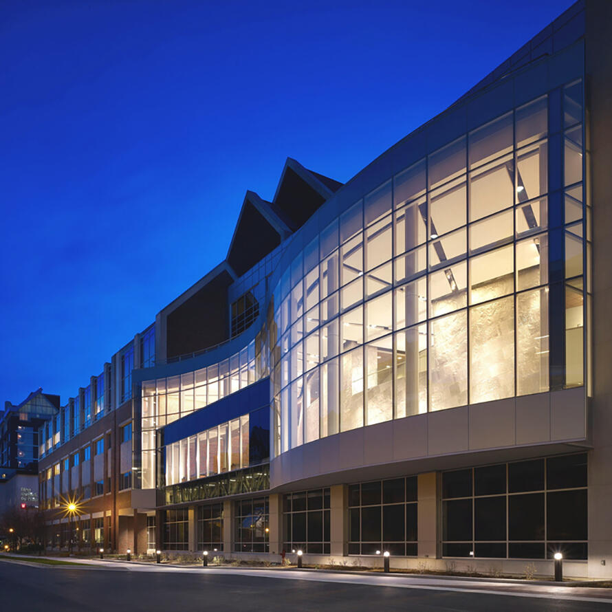 Indiana University Melvin and Bren Simon Cancer Center with a curved glass facade illuminated at dusk along a quiet street.