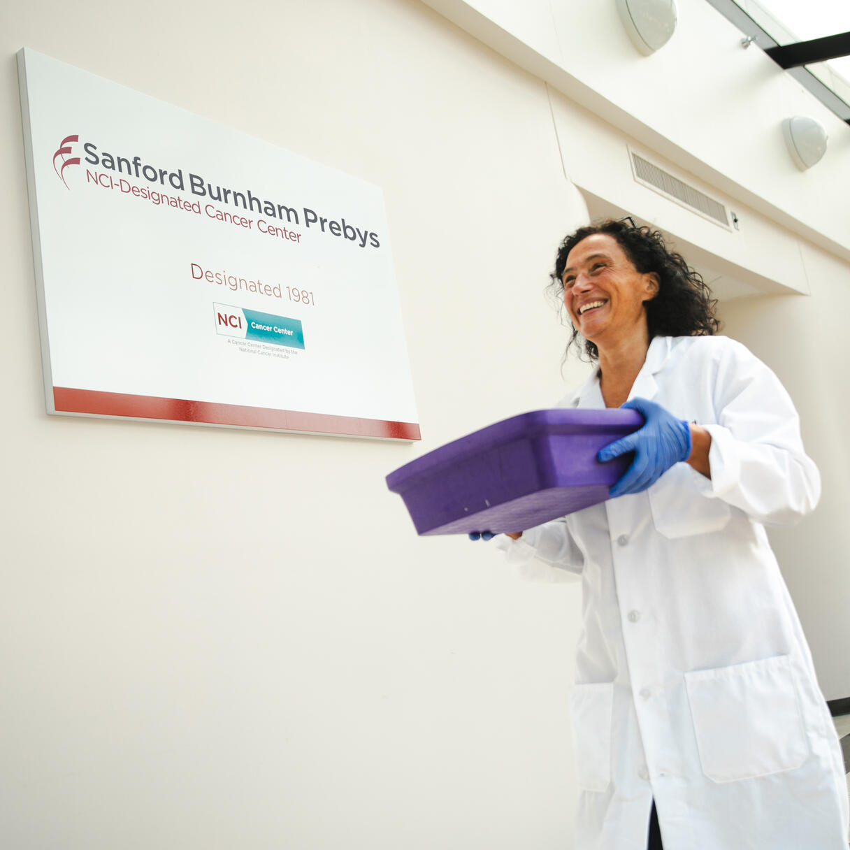 Person in a lab coat carrying a tray in a bright hallway at Sanford Burnham Prebys Medical Discovery Institute beside a wall sign.
