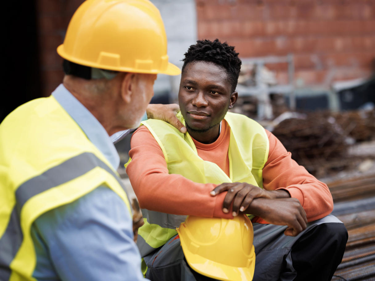 Dos obreros con vestimenta de construcción conversan mientras uno de ellos apoya una mano sobre el hombro del otro.