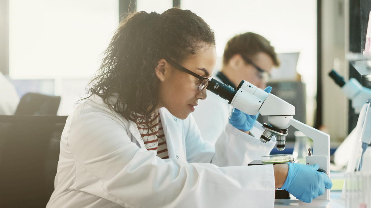 A young, female scientist in a lab coat and gloves examines a sample under a microscope in a laboratory, while two colleagues work in the background.